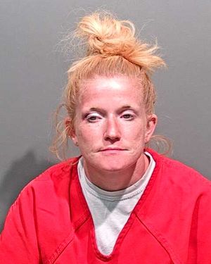 A woman with blonde hair styled in a bun wears a red prison jumpsuit and has a neutral expression.