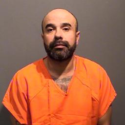 A man with a beard wearing an orange prison jumpsuit is facing the camera.