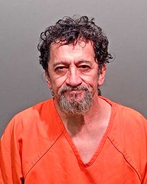 A middle-aged man with curly hair and a beard is smiling while wearing an orange prison jumpsuit against a gray background.