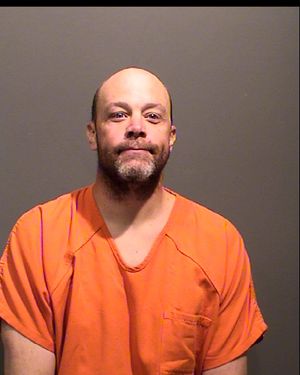 A man wearing an orange prison jumpsuit poses for a mugshot against a gray background.