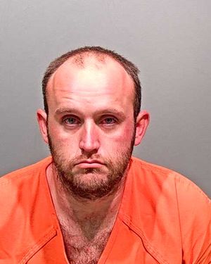 A man with a shaved head and beard is shown wearing an orange prison jumpsuit against a gray background.