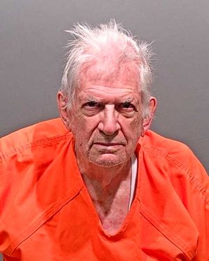 An elderly man with disheveled white hair is wearing an orange jumpsuit and looking directly at the camera.