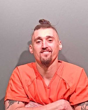 A man with a unique hairstyle and tattoos is smiling while wearing an orange jumpsuit against a gray background.