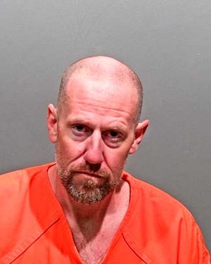 A man with a shaved head and beard is shown wearing an orange prison jumpsuit against a gray background.