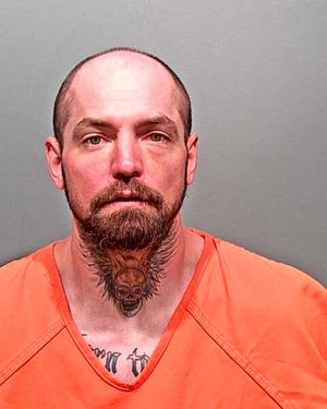 A man with a shaved head and a prominent beard, wearing an orange prison jumpsuit, poses for a mugshot against a plain background.