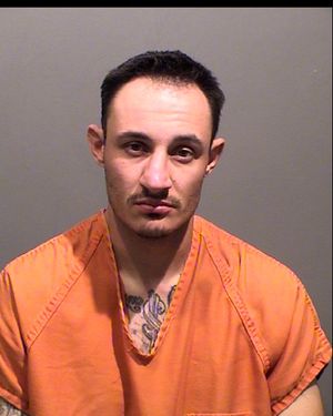 A man in an orange jumpsuit poses for a mugshot, displaying tattoos on his chest and a stern expression.