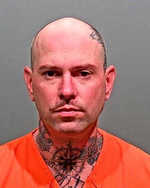 A bald man with tattoos on his neck and chest is wearing an orange inmate jumpsuit and looking directly at the camera.