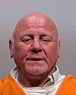 A man with a shaved head, wearing an orange prison jumpsuit, poses for a mugshot against a plain background.