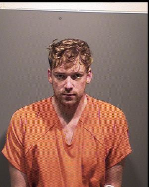 A young man with tousled hair is wearing an orange prison jumpsuit and looking directly at the camera against a plain gray wall.
