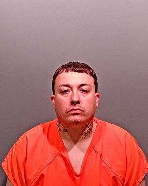 A man with tattoos wearing an orange jumpsuit poses for a mugshot against a gray background.