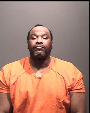 A man wearing an orange prison jumpsuit stands against a plain background.