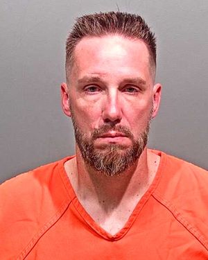 A man with a beard and short hair is wearing an orange prison jumpsuit and looking directly at the camera.