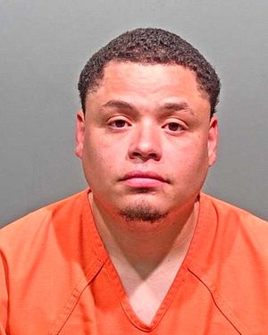 A man in an orange prison jumpsuit poses for a mugshot against a plain background.