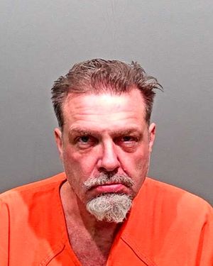 A man with a serious expression is wearing an orange jail jumpsuit against a gray background.