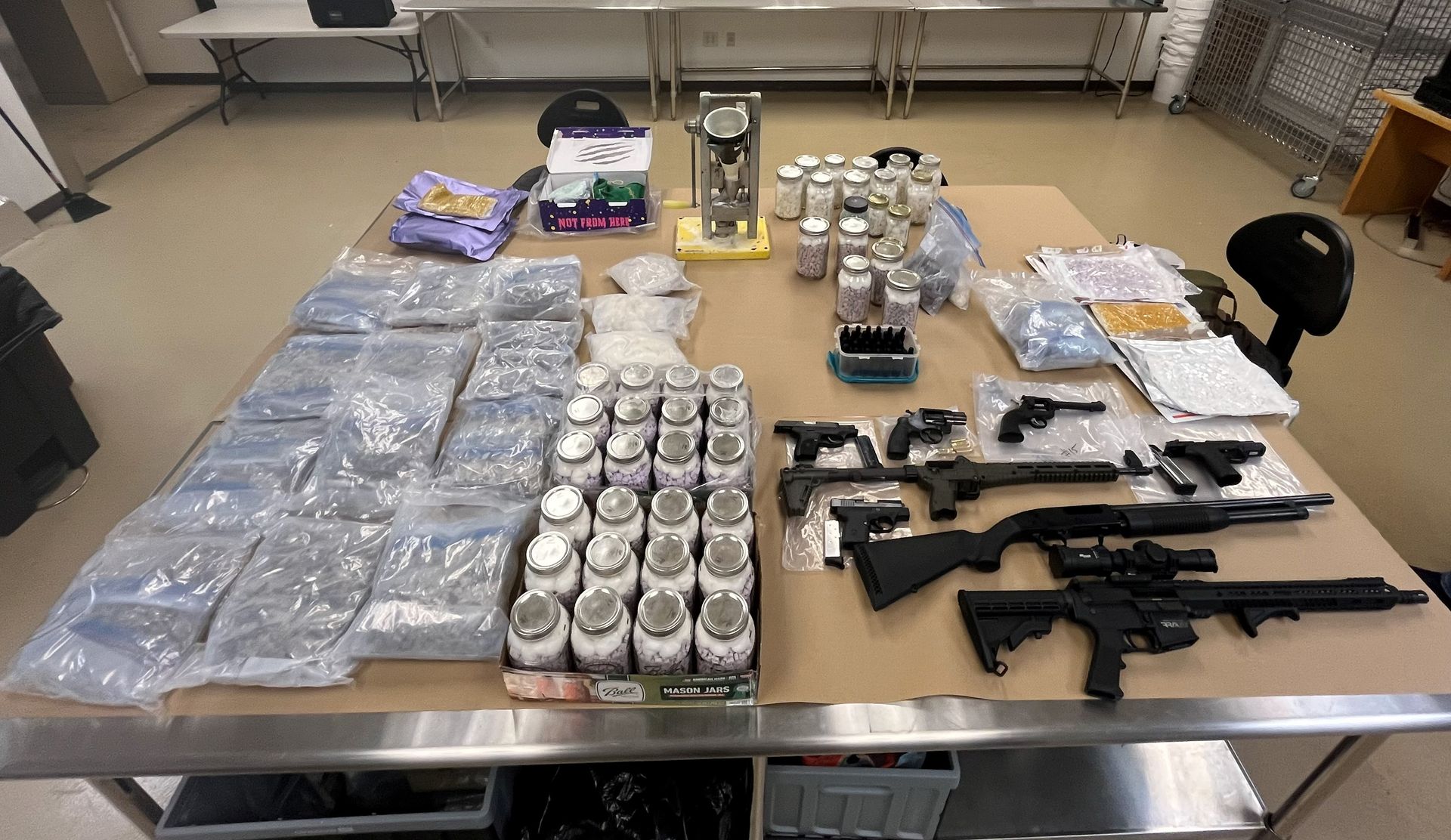 Evidence displayed on a large table in a processing room, including multiple sealed bags, jars containing pills or suspected narcotics, several handguns, two rifles, a shotgun, and drug-packaging equipment.