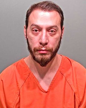 A man with a beard wearing an orange prison jumpsuit is looking directly at the camera.
