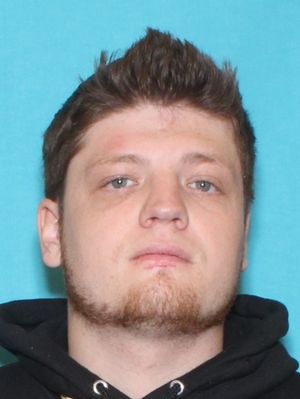 A young man with light brown hair and a beard is shown against a blue background, wearing a black hoodie.