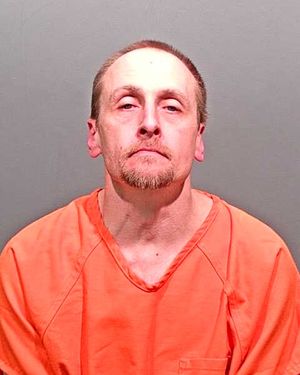 A man wearing an orange prison jumpsuit is posed for a mugshot against a gray background.